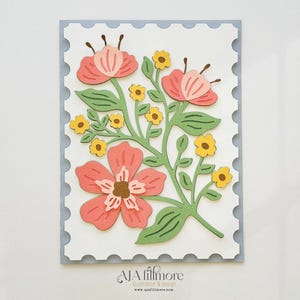 May include: A white card with a blue border and a floral design. The flowers are pink, yellow, and green. The card has the text "AJA fillmore" at the bottom.