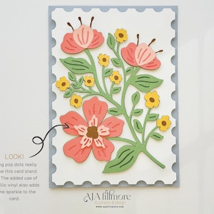 May include: A white card with a scalloped border features a floral design with pink and yellow flowers and green leaves. The text "LOOK! Using pop dots really make this card stand out. The added use of metallic vinyl also adds some sparkle to the card." is printed below the floral design. The text "JA filfmore Illustration & Design www.jafilfmore.com" is printed at the bottom of the card.