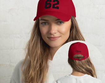 May include: A red baseball cap featuring the black embroidered number 62 on the front crown. The back of the cap includes an adjustable metal buckle strap and the black embroidered name Chris centered above the opening.