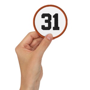 May include: A circular embroidered patch featuring a white background with the black number 31 centered in a bold font. The patch is finished with a thick, burnt orange stitched border that frames the entire circular design clearly.