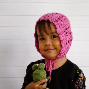 May include: A young child wearing a pink crocheted bonnet against a white background. The child is holding a green crocheted frog toy.