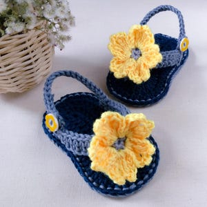 May include: A pair of crocheted baby sandals with yellow flower embellishments. The sandals are blue and gray with yellow button accents.