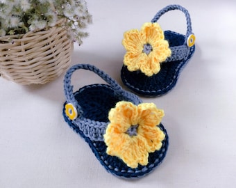May include: A pair of crocheted baby sandals with yellow flower embellishments. The sandals are blue and grey with yellow button accents.