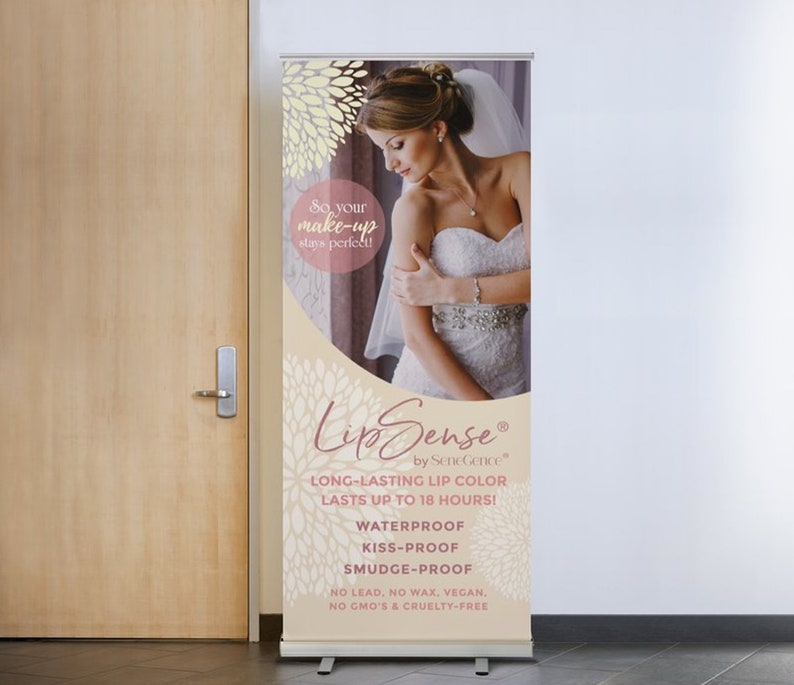 May include: A white retractable banner with a pink and white floral design. The banner advertises Lip Sense long-lasting lip color that lasts up to 18 hours. The banner also states that the lip color is waterproof, kiss-proof, and smudge-proof. It is also vegan, cruelty-free, and contains no lead, wax, or GMOs.