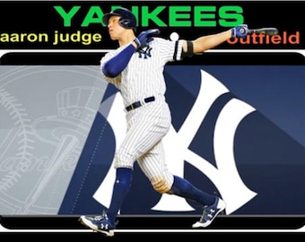 May include: A baseball player wearing a white and blue uniform with the word "YANKEES" in green letters on the front of the jersey. The player is swinging a bat and the text "aaron judge" and "outfield" are visible in the image.