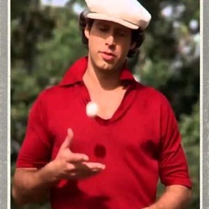 May include: A trading card featuring a person wearing a red polo shirt and a cream-colored flat cap, juggling a white golf ball. The bottom of the card displays the text Ty Webb and BUSHWOOD in a gray border with a white frame.