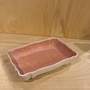 May include: A rectangular ceramic dish with a wavy, scalloped rim. The interior of the dish features a smooth, matte dusty rose pink glaze, while the exterior walls are finished in a contrasting, glossy off-white glaze with visible texture.