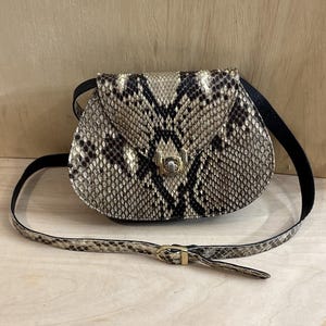 May include: A crossbody bag featuring a beige and black snakeskin pattern with a rounded flap closure. The bag includes a gold-toned metal turn-lock clasp and a matching adjustable shoulder strap with a gold-toned buckle.