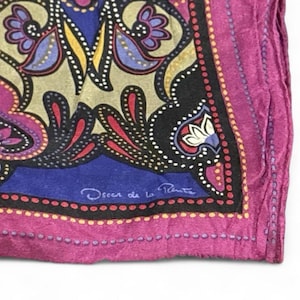 May include: A square scarf featuring a vibrant paisley pattern in shades of magenta, royal blue, black, and cream. The design includes intricate floral motifs and a dotted border. The signature Oscar de la Renta is printed in white on the blue section.