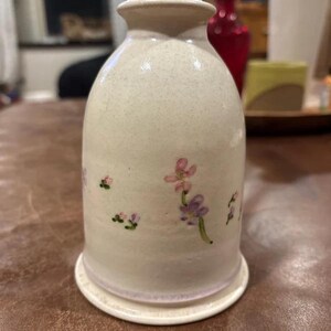 Puede incluir: Un jarrón de cerámica de color crema con un acabado moteado y un cuello estrecho. El cuerpo del jarrón está decorado con pequeños motivos florales pintados a mano en tonos rosa y morado, con delicados tallos y hojas verdes sobre la superficie.