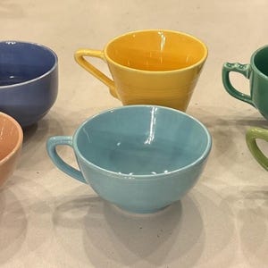 Op de afbeelding: Een set van zes keramische kopjes in diverse effen kleuren, waaronder perzik, blauw, geel, turquoise, groen en limoengroen. Elk kopje heeft een gladde, glanzende afwerking, een ronde komvorm en een enkel handvat aan de zijkant om vast te houden.