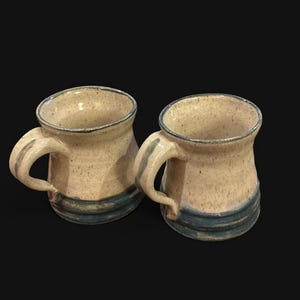 Może przedstawiać: Dwa ceramiczne kubki z nakrapianą kremową glazurą i ciemnoniebieskim pasem u podstawy. Każdy kubek posiada solidne ucho oraz rozszerzany ku górze brzeg, co nadaje im ręcznie wykonany wygląd z subtelnymi różnicami w nakrapianej fakturze.