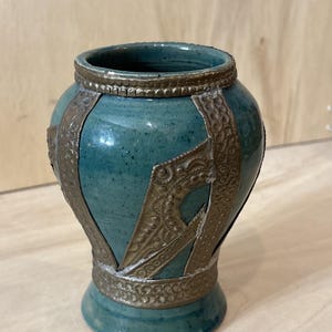 May include: A teal ceramic vase with a glossy, speckled finish, featuring decorative bronze-colored metal bands around the rim, base, and vertical sections. The metal accents display intricate, embossed patterns throughout the surface.