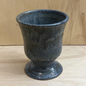 May include: A ceramic goblet with a flared rim and a pedestal base. The exterior features a mottled glaze in shades of dark blue, grey, and brown, creating a textured appearance with subtle variations in color across the surface of the vessel.