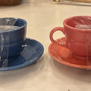Könnte beinhalten: Zwei Keramik-Teetassen mit passenden Untertassen, eine in Dunkelblau und die andere in Korallenrosa. Jede Tasse hat einen abgerundeten Henkel und horizontale Rillen an der Außenseite. Beide Sets sind in durchsichtige Schutzfolie eingewickelt.