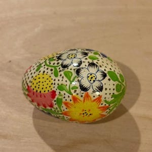 May include: An egg-shaped object with a cream-coloured base featuring a hand-painted floral pattern. The design includes black-outlined white flowers, a bright yellow sunflower, a pink flower, and green leaves, all set against a background of small black dots.