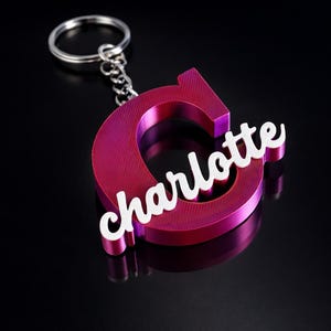 Puede incluir: Un llavero magenta con la letra "C" y el nombre "Charlotte" en escritura blanca. Tiene una cadena y un anillo plateados. El diseño está sobre un fondo negro.
