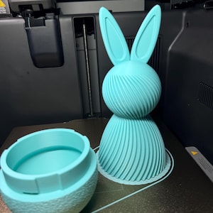 Puede incluir: Una figura de conejo de Pascua impresa en 3D de color azul claro con un recipiente a juego. El conejo tiene orejas largas y un diseño en espiral en su cuerpo. El recipiente tiene un exterior texturizado y un interior liso.