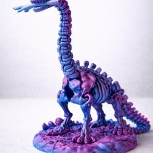 May include: A purple and blue dinosaur skeleton figurine with a long neck and tail. The figurine is standing on a round base. The dinosaur's tongue is sticking out. The color scheme is a gradient of purple and blue.