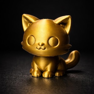 Op de afbeelding: Een gouden kattenbeeldje met grote, ronde ogen en een kleine, lachende mond. De kat zit met opgerolde staart. Het beeldje is gemaakt van een glanzend, metallic materiaal tegen een donkere achtergrond.