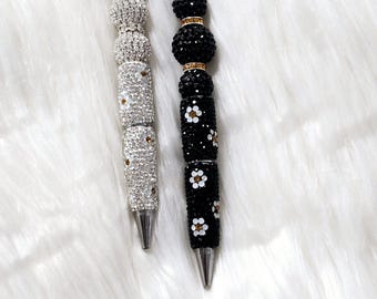 May include: Two decorative pens with jeweled accents. One pen is silver with clear rhinestones, and the other is black with white daisy accents. Both pens have a silver tip and are on a white, fluffy surface.