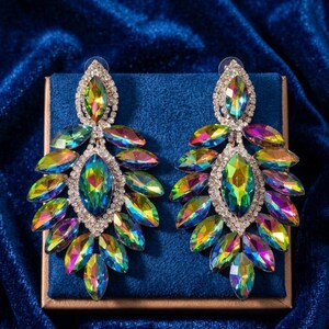 Puede incluir: Un par de pendientes tipo candelabro con cristales iridiscentes de corte marquesa en tonos verde, azul y morado. Los cristales están dispuestos en forma de hoja y bordeados por pequeños brillantes transparentes en una montura de metal plateado.