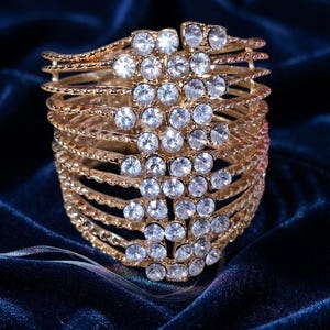 May include: A wide gold-tone cuff bracelet featuring multiple horizontal textured bands connected by a vertical central panel encrusted with numerous clear, round, faceted rhinestones that catch the light, creating a brilliant, reflective appearance.