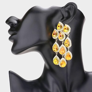 May include: A chandelier earring featuring multiple teardrop-shaped yellow crystals arranged in a cascading pattern. Each crystal is framed by small clear stones set in a gold-toned metal base, creating a tiered, dangling design that hangs vertically.