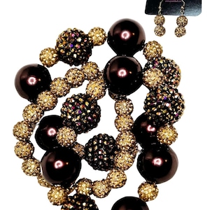 Puede incluir: Un collar y un juego de pendientes a juego. El collar presenta grandes cuentas de color marrón oscuro intercaladas con cuentas más pequeñas de color dorado cubiertas de cristal. Los pendientes tienen un diseño similar. La marca "J Cardock" es visible.
