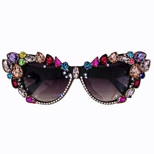 May include: Cat-eye sunglasses featuring dark tinted lenses and a black frame. The outer edges and bridge are heavily embellished with an assortment of multi-colored, faceted crystals in shades of purple, pink, blue, green, and clear white.