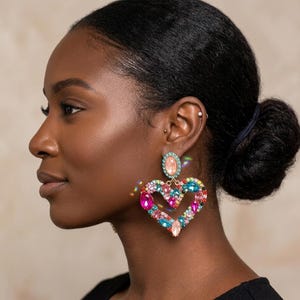 May include: A large heart-shaped dangle earring featuring a central oval peach-colored stone at the top, connected to a multi-colored heart frame encrusted with various faceted crystals in shades of pink, blue, orange, and clear, set in gold-tone metal.