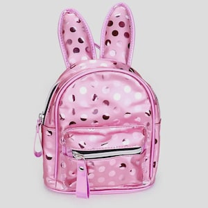 Könnte beinhalten: Ein glänzender, metallisch rosa Rucksack mit einem Punktemuster und zwei aufrechten Hasenohren an der Oberseite. Die Tasche verfügt über ein Hauptfach mit Reißverschluss und eine kleinere Vordertasche mit einem langen rosa Reißverschlussanhänger.
