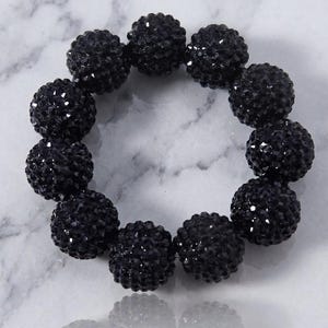May include: A circular bracelet composed of eleven spherical beads with a faceted, textured surface. Each bead is a deep, solid black colour with a reflective, shimmering finish that catches the light across its multiple small, geometric facets.