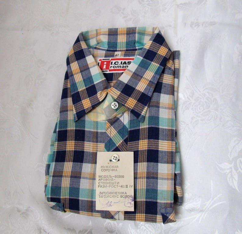 May include: A blue, yellow, and green plaid shirt with a white collar. The shirt has a label that reads "I.C.IAS roman" and "МУЖ CHAR СОРОЧНА МОДЕЛЬ-В0300 АРТИКУЛ- СТОЕНЕШТИ РОСТ-42 РАЗМ-РОСТ-42 IV 50% СИНТЕТИКА 50%ИСКУС ВОЛОК 12".