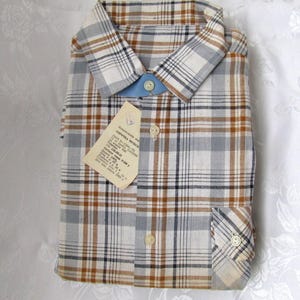 May include: A plaid shirt with a brown, blue, and white pattern. The shirt has a button-down collar and a pocket on the left side. The shirt has a tag attached to it with the text "CONFEZIONE ARTICOLO" in Italian.