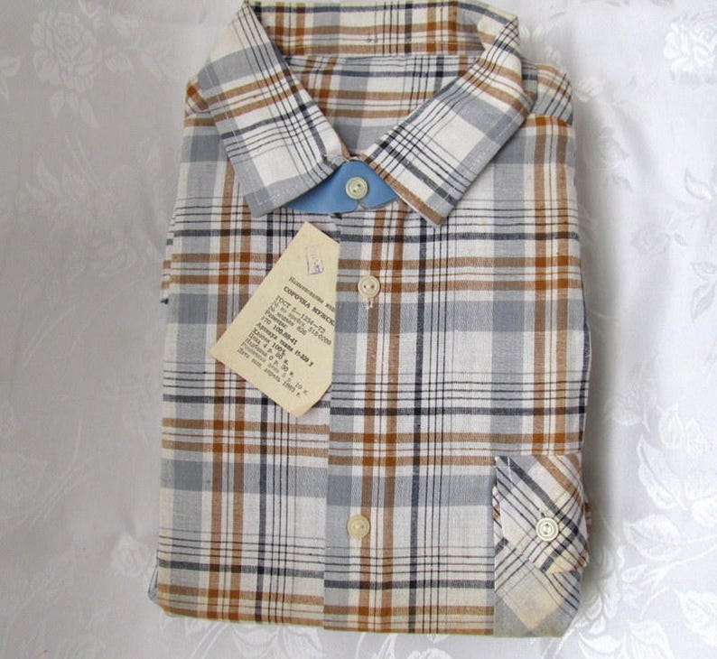 May include: A plaid shirt with a brown, blue, and white pattern. The shirt has a button-down collar and a pocket on the left side. The shirt has a tag attached to it with the text "CONFEZIONE ARTICOLO" in Italian.