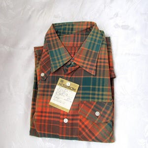 May include: A plaid shirt with a green, orange, and brown color scheme. The shirt has a button-down collar and a pocket on the left side. The shirt has a tag attached that says "Каштан".