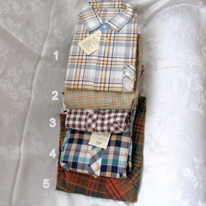 May include: A stack of five plaid shirts in different colors and patterns. The shirts are folded and stacked on top of each other. The top shirt is white with brown and blue plaid, the second shirt is green with brown plaid, the third shirt is brown and white checkered, the fourth shirt is blue and white plaid, and the bottom shirt is orange and brown plaid.