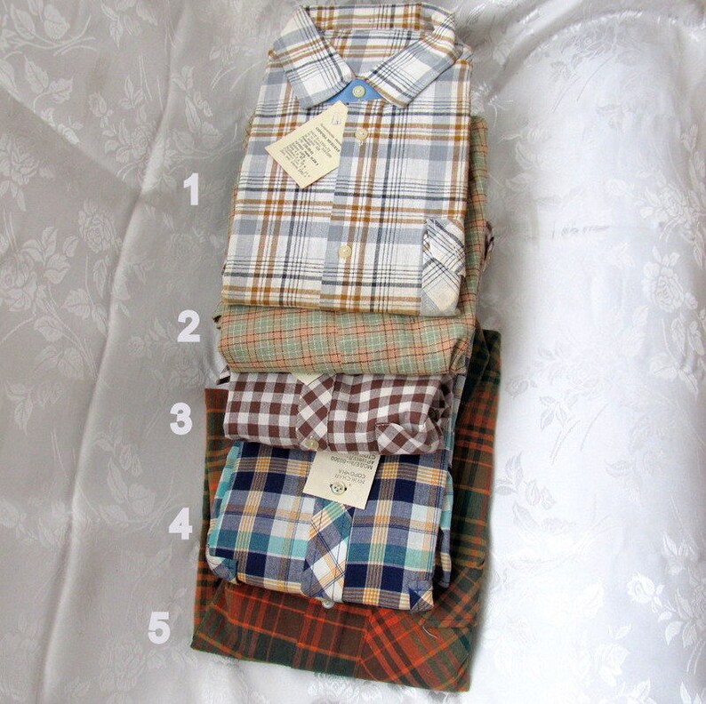 May include: A stack of five plaid shirts in different colors and patterns. The shirts are folded and stacked on top of each other. The top shirt is white with brown and blue plaid, the second shirt is green with brown plaid, the third shirt is brown and white checkered, the fourth shirt is blue and white plaid, and the bottom shirt is orange and brown plaid.