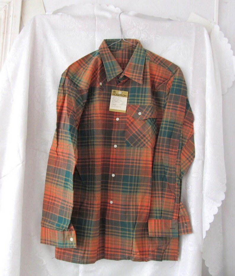 May include: A long-sleeved plaid shirt with a button-down collar. The shirt is orange, green, and brown. The shirt has a pocket on the left side and a tag that says "NOVITCH" on the front.
