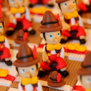 May include: A collection of small, hand-crafted figurines of Pinocchio. Each figurine is dressed in a red and white outfit with a brown hat and a long, wooden nose. The figurines are arranged on a brown surface.