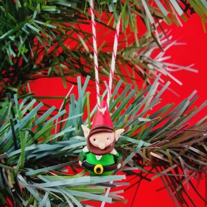 Peut inclure: Une décoration de gnome verte et rouge avec une barbe blanche et une boucle de ceinture dorée suspendue à une ficelle rouge et blanche sur un sapin de Noël vert.