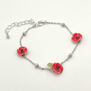 Könnte beinhalten: Silberfarbenes Armband mit drei roten Mohnblumen-Anhängern mit schwarzen Zentren und einem grünen Blatt. Das Armband hat ein Ketten-Design mit kleinen silbernen Perlen und einem verstellbaren Verschluss.