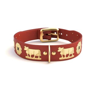 Op de afbeelding: Een rode leren hondenhalsband met goudkleurige metalen accenten. De halsband heeft gouden koe- en bloemmotieven, een gouden gesp en een gouden lus. De halsband is ontworpen voor een huisdier.