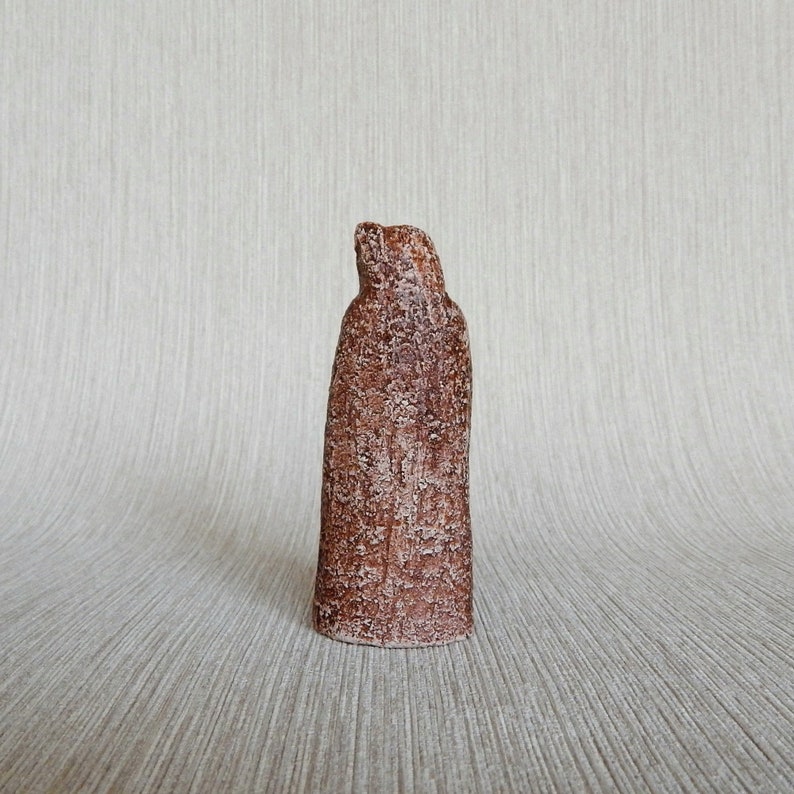 May include: A brown ceramic sculpture of a person wearing a long robe. The sculpture has a rough, textured surface.