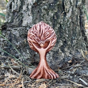 May include: A brown clay sculpture of a tree with a human-like figure integrated into the trunk. The branches extend upwards and outwards, forming a canopy of leaves. The base of the sculpture features textured roots that flare out at the bottom.