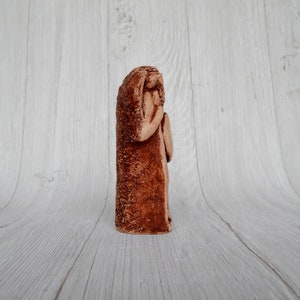 May include: A terracotta sculpture of a person with their head bowed, appearing to be in prayer or contemplation. The sculpture is a reddish-brown color and has a rough, textured surface.
