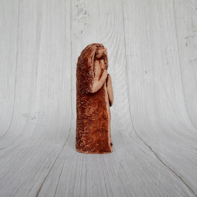 May include: A terracotta sculpture of a person with their head bowed, appearing to be in prayer or contemplation. The sculpture is a reddish-brown color and has a rough, textured surface.