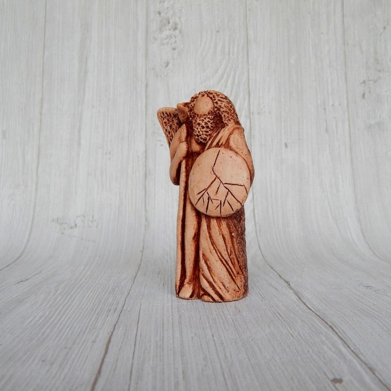 May include: A terracotta figurine of a person holding a shield with a tree design. The figurine is standing on a white wooden surface.