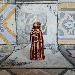 May include: A brown ceramic figurine of a person wearing a long robe and holding a lyre. The figurine is standing on a tiled surface.